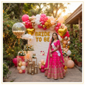 Punjabi Phulkari Bride decoration with pink, peach and gold balloon arch, gold heart foil balloons, Bride To Be foil letters and elegant outdoor bridal shower backdrop.